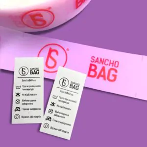 Рожева стрічка та дві білі тканинні етикетки Sancho Bag.